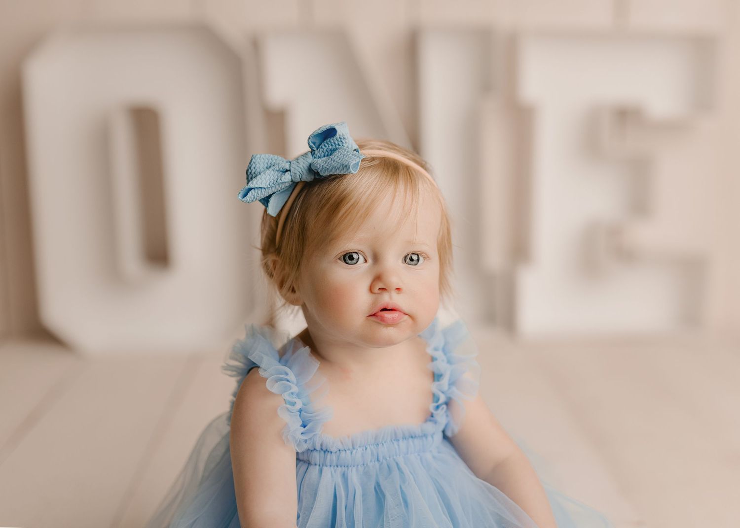 A baby girl in a blue tutu dress with a bow headband sits in front of large white ONE letters for her first birthday.
