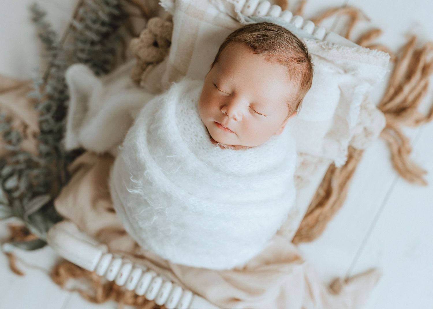 Newborn baby swaddled in white wrap sleeping peacefully in a rustic basket with dried botanicals.