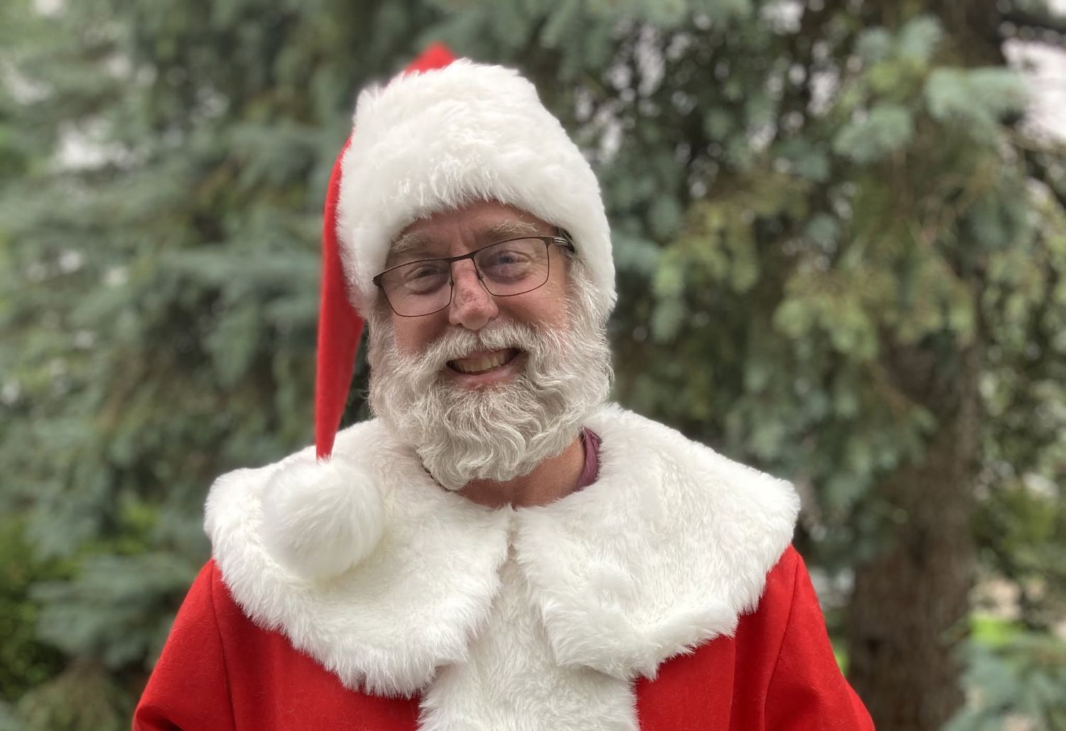 Smiling man dressed as Santa Claus in red suit with white fur trim and hat, wearing glasses outdoors.
