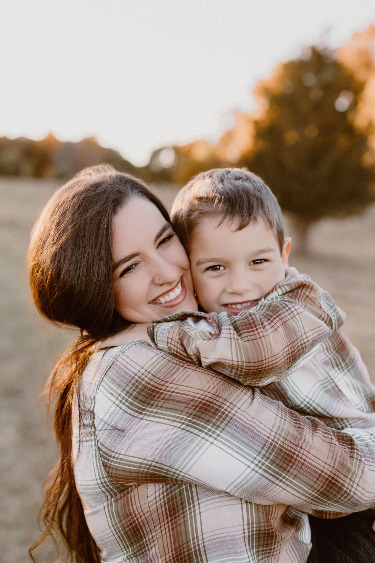 A smiling young woman hugs a happy toddler boy outdoors during golden hour in a sunlit autumn field.