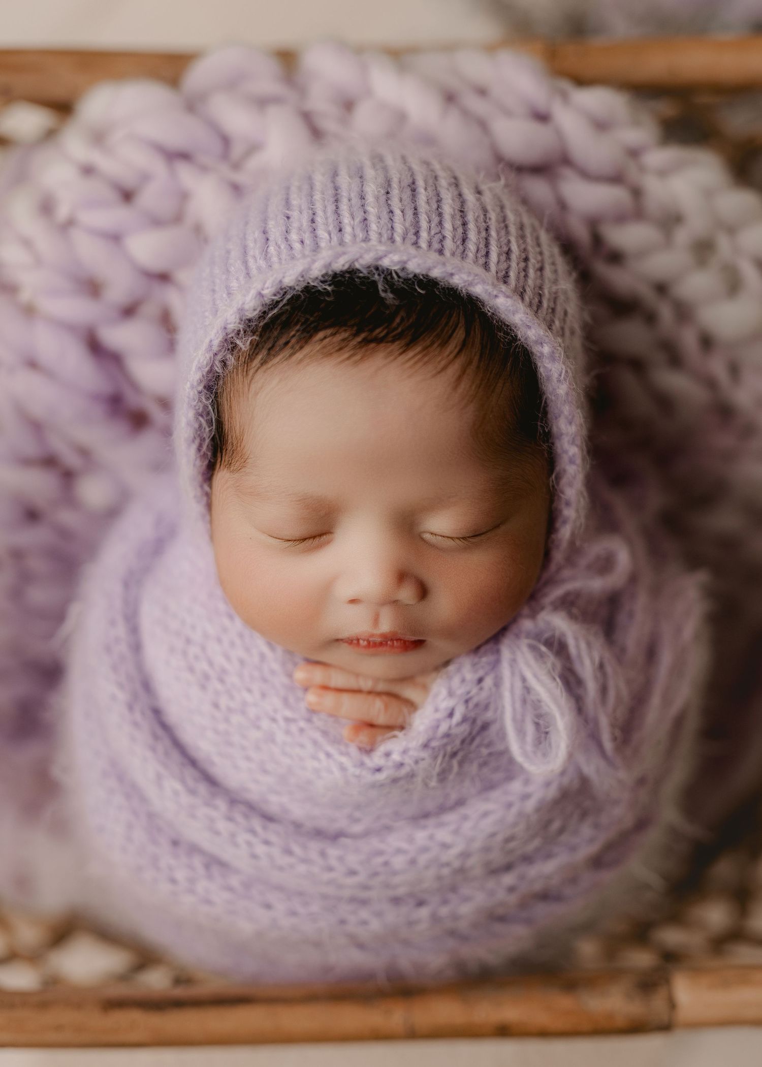 Newborn baby sleeping peacefully wrapped in a soft lavender knit blanket and matching bonnet hat.