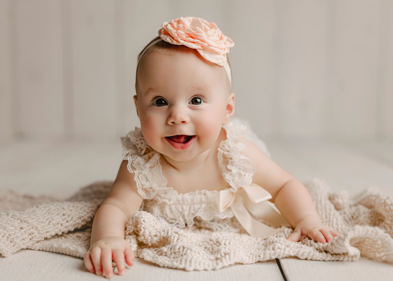 Smiling baby girl in cream lace outfit and peach flower headband lying on knitted blanket.