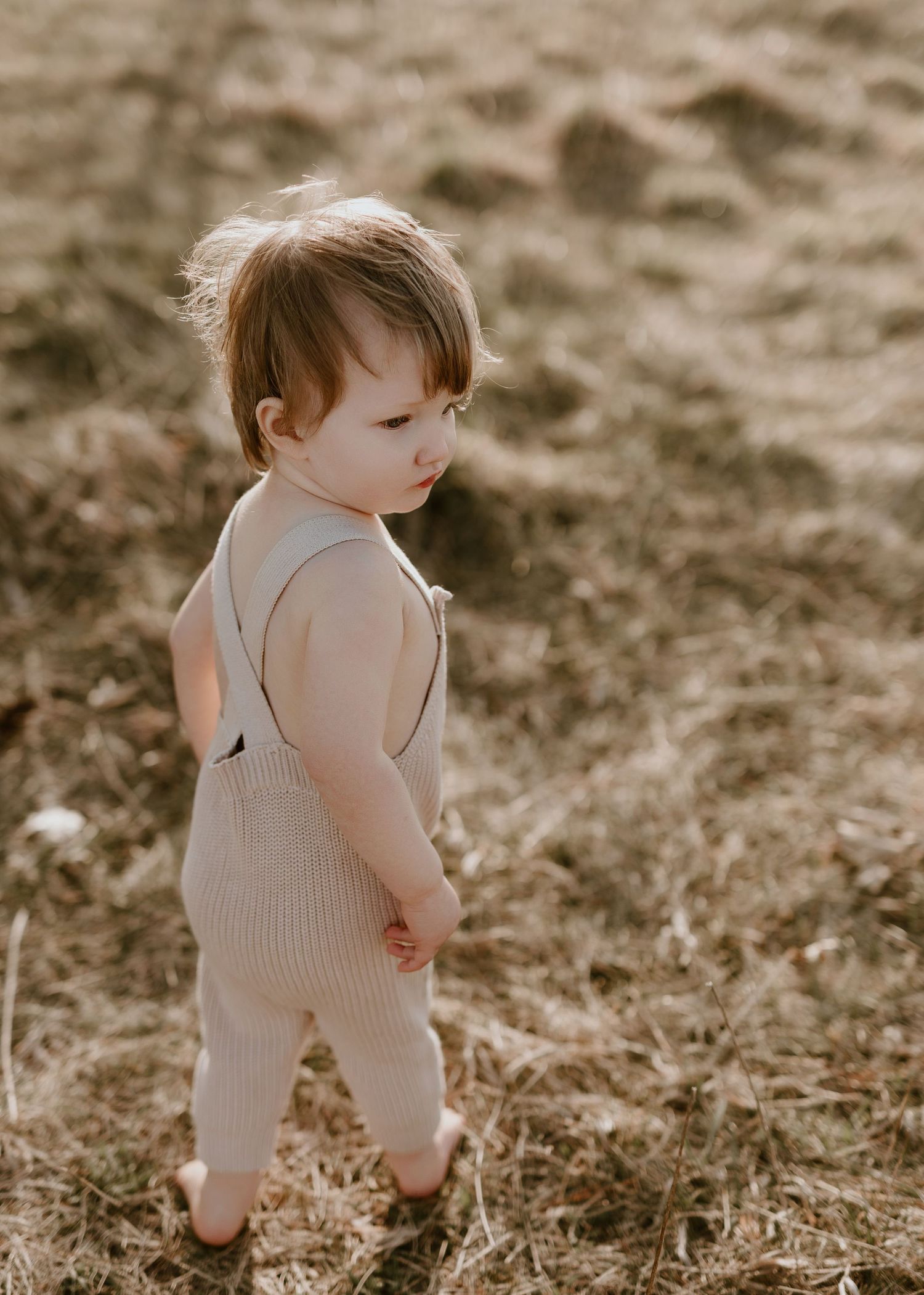 Toddler in a beige knit romper standing barefoot on dry grass in a sunlit field.