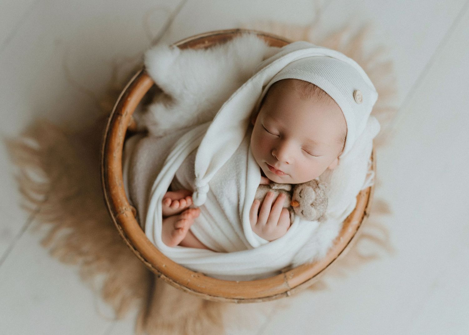 Newborn baby swaddled in white wrap sleeping peacefully in a wooden bowl with soft fur, wearing a knit bonnet.
