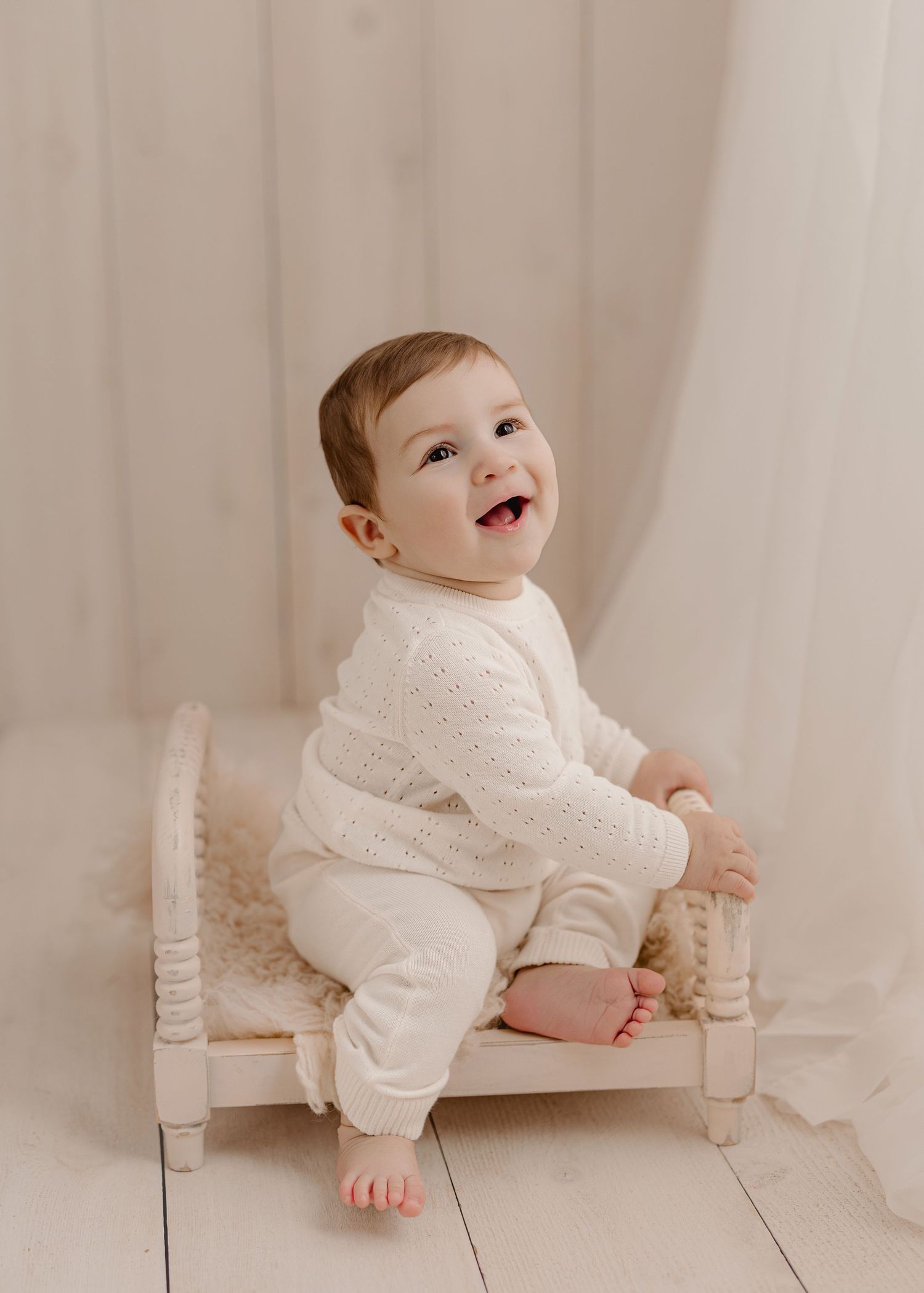Smiling baby in cream knit outfit sitting on a miniature white wooden bed against a soft white backdrop.