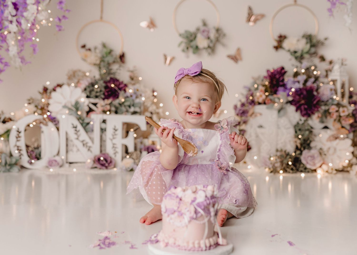 Baby girl in purple dress smiles at her first birthday cake smash photo session with floral backdrop.