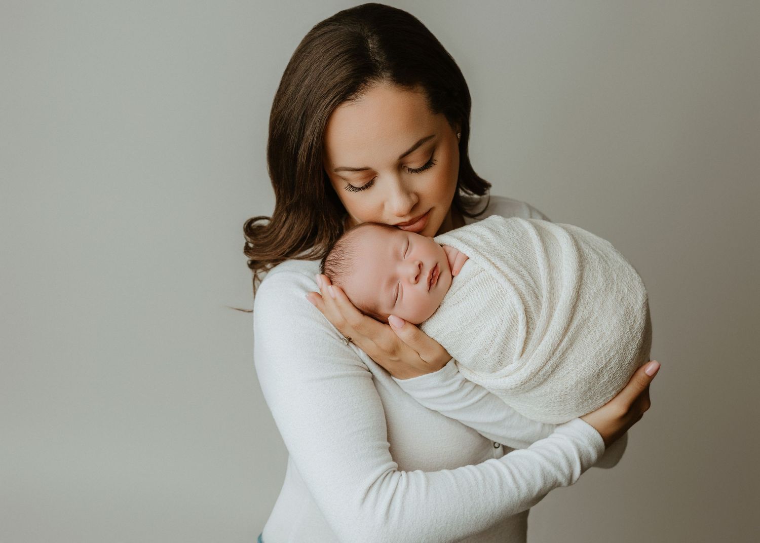 A mother in a white dress tenderly holds her swaddled newborn baby close to her chest against a soft gray background.