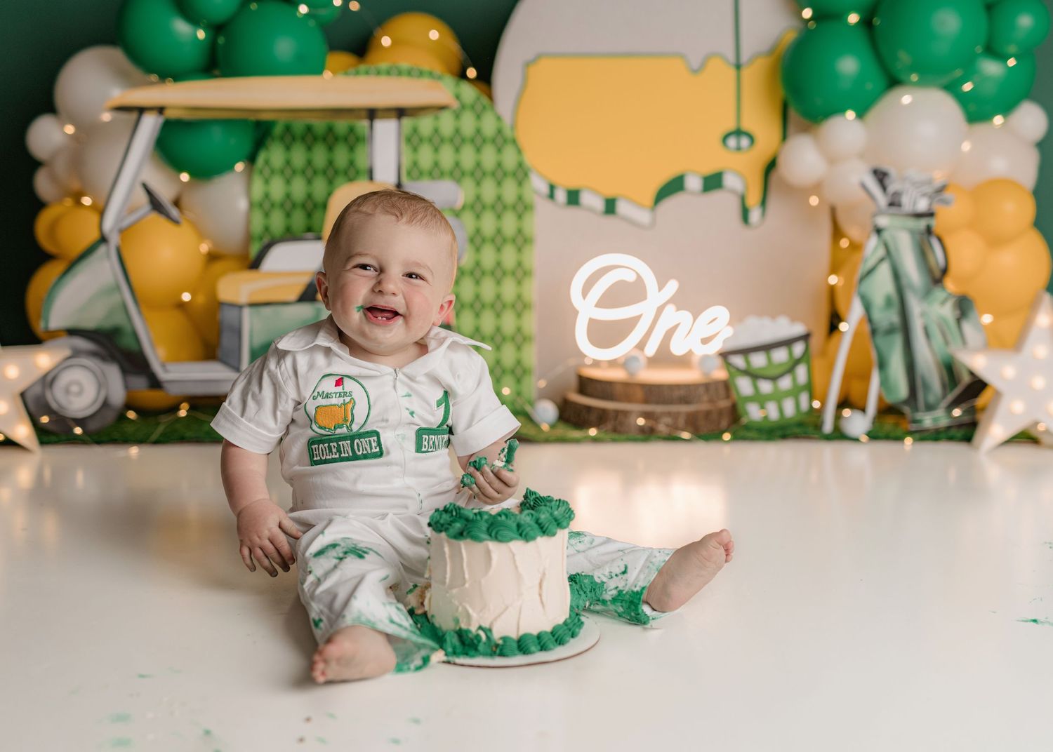 Baby boy smiles at his golf-themed first birthday cake smash with green balloons and a golf cart backdrop.
