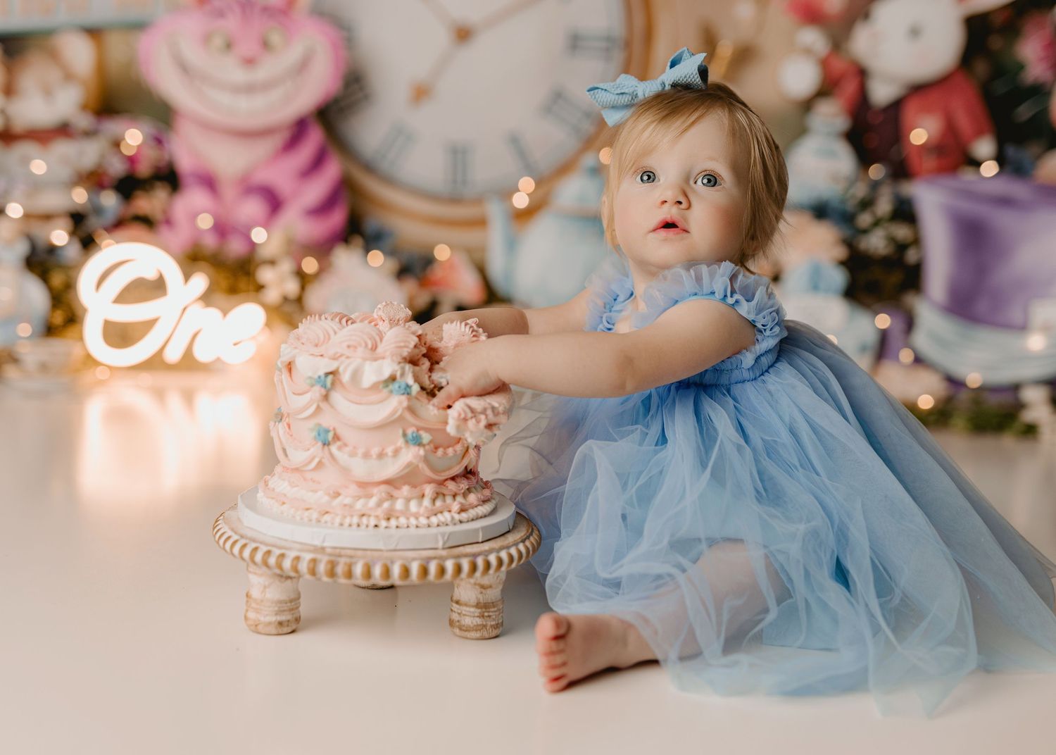 Baby girl in blue tulle dress touches pink cake at Alice in Wonderland themed first birthday smash cake session.