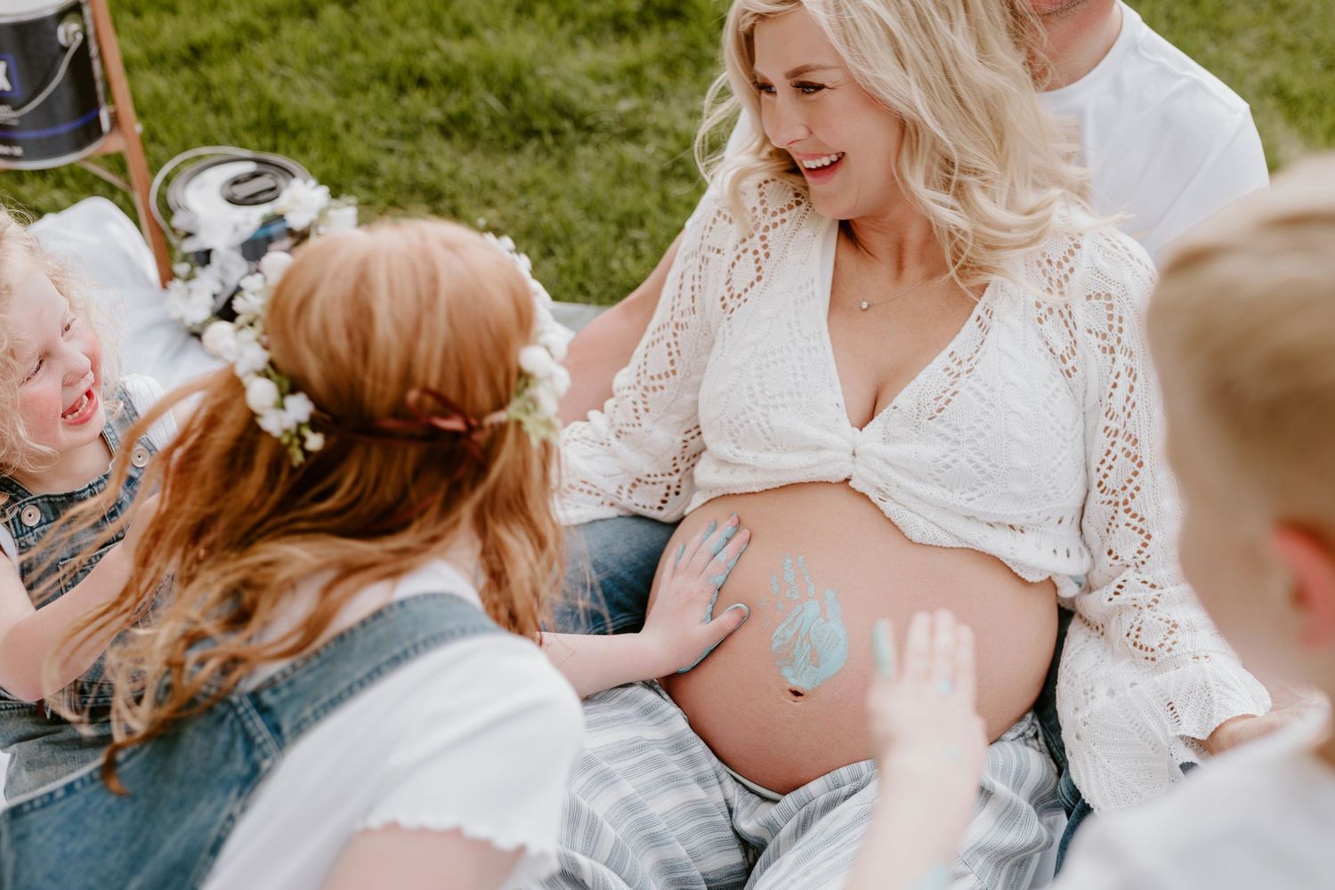 Pregnant woman in white top sits outdoors as children touch her painted baby bump during a maternity photo session in Oakland County, Mi