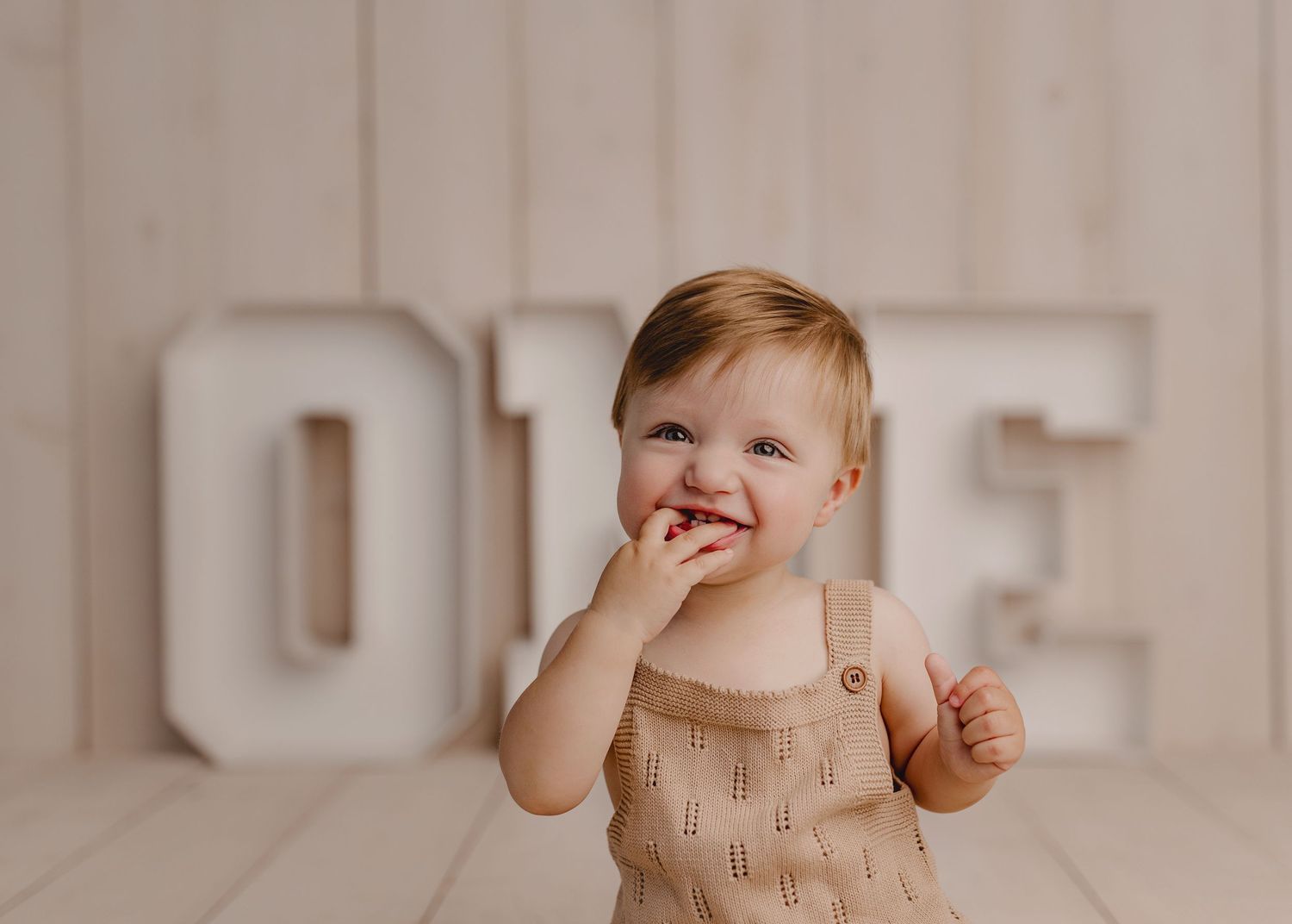 Smiling toddler in beige romper eating cake in front of large ONE letters for first birthday photo.