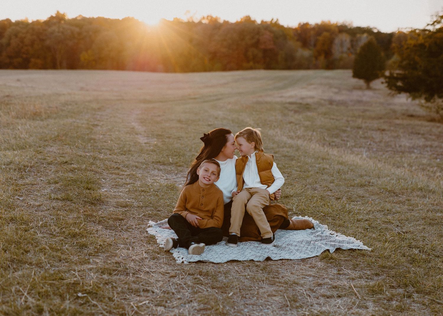 A mother sits on a blanket in a golden field at sunset, embracing her two young children warmly.