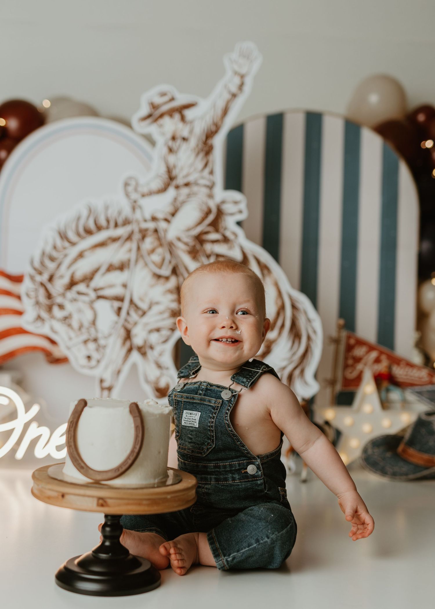 Baby boy in denim overalls smiles at his first birthday cake smash with a western cowboy themed backdrop.