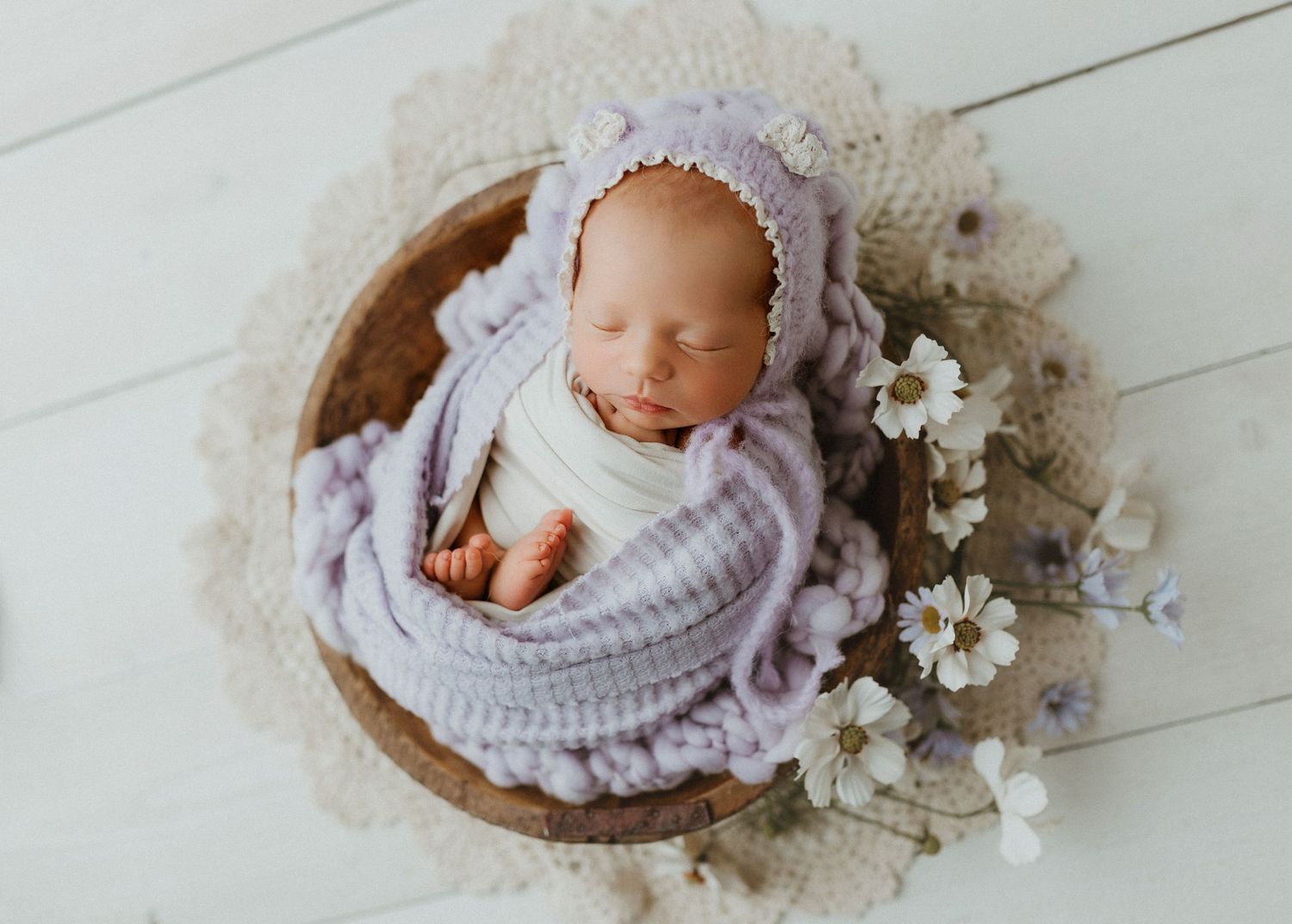 Newborn baby wrapped in lavender knit blanket and bonnet, sleeping in wooden bowl with white flowers.