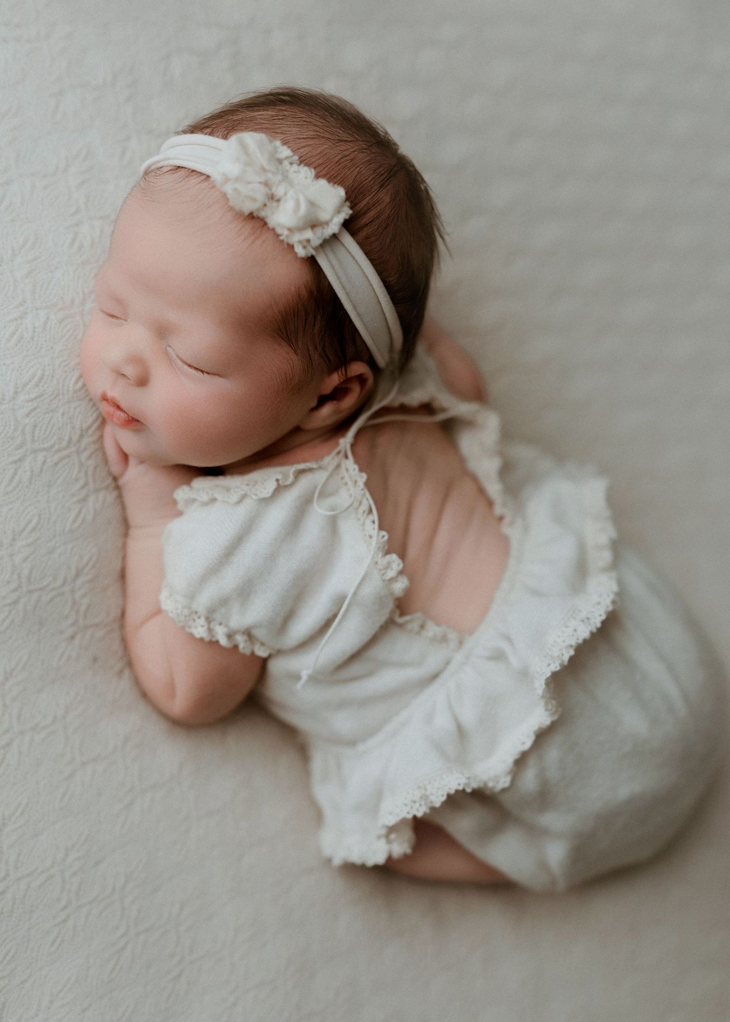 Sleeping newborn baby girl in white romper and floral headband, posed on cream textured blanket.