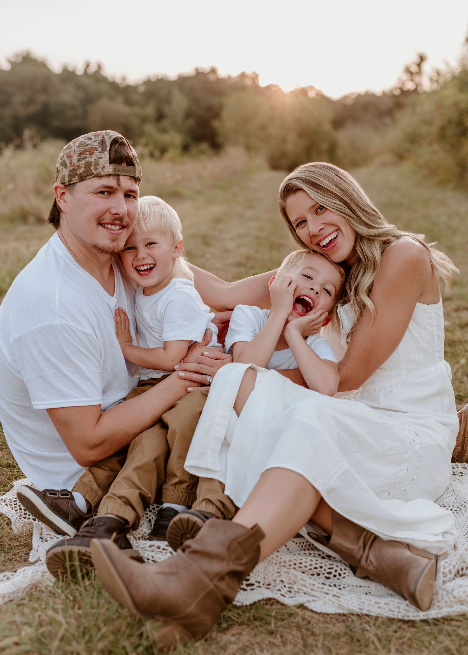 Happy family of four sitting on blanket in golden hour field, parents laughing with two young children in white outfits.