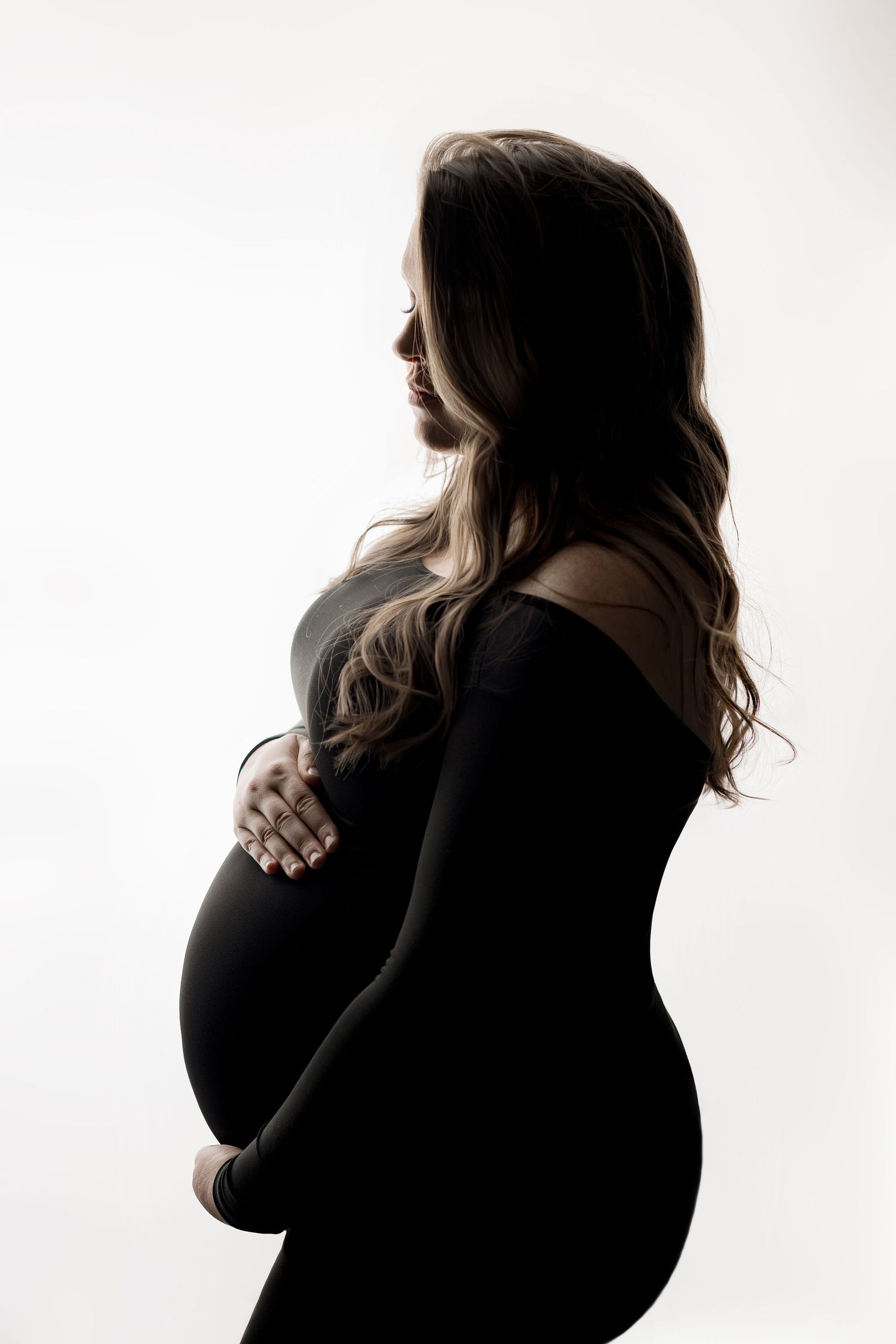 Silhouette of a pregnant woman in a fitted black off-the-shoulder gown cradling her baby bump against a bright white background.