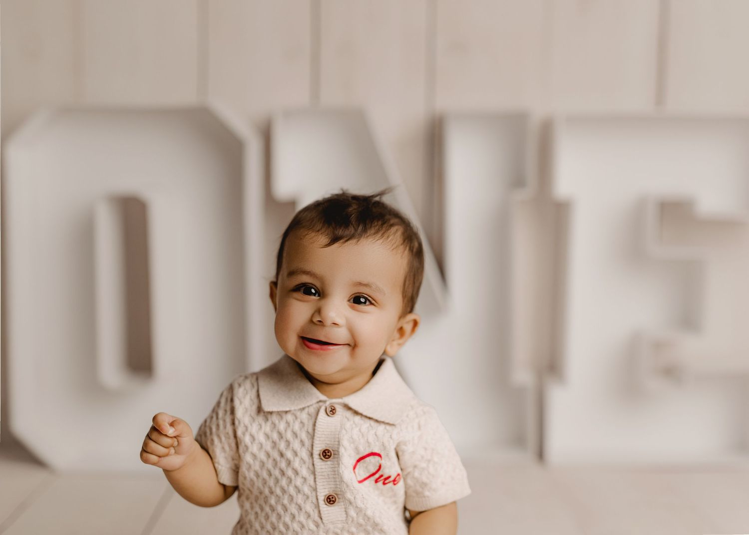 Smiling baby in cream knit romper sitting before large ONE letters for first birthday photo.