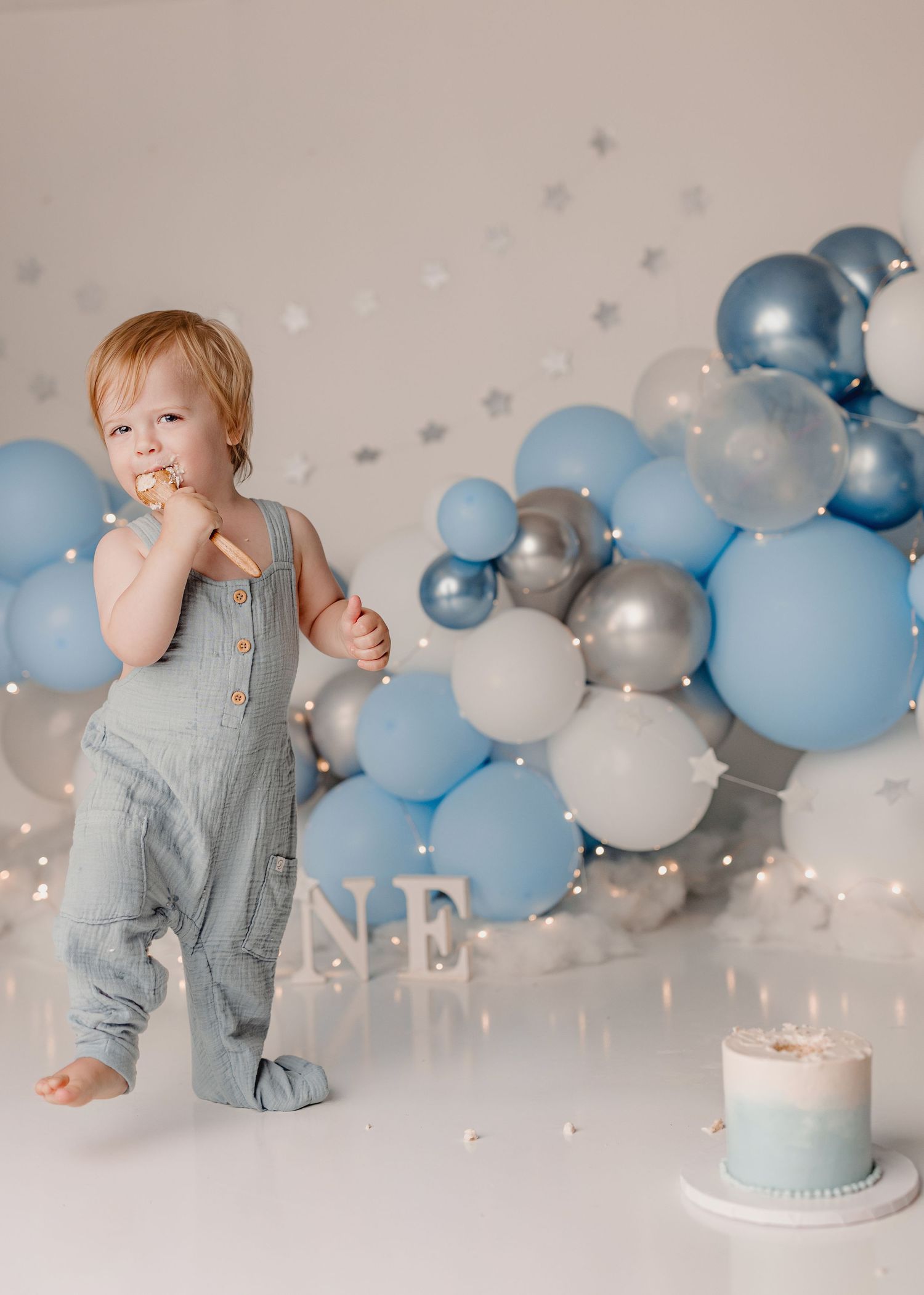 Toddler boy in blue overalls eating cake at a first birthday party with blue and silver balloon backdrop.