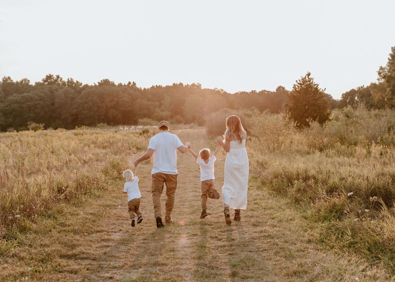 Family of four walking hand-in-hand down a sunlit dirt path through golden fields at sunset.