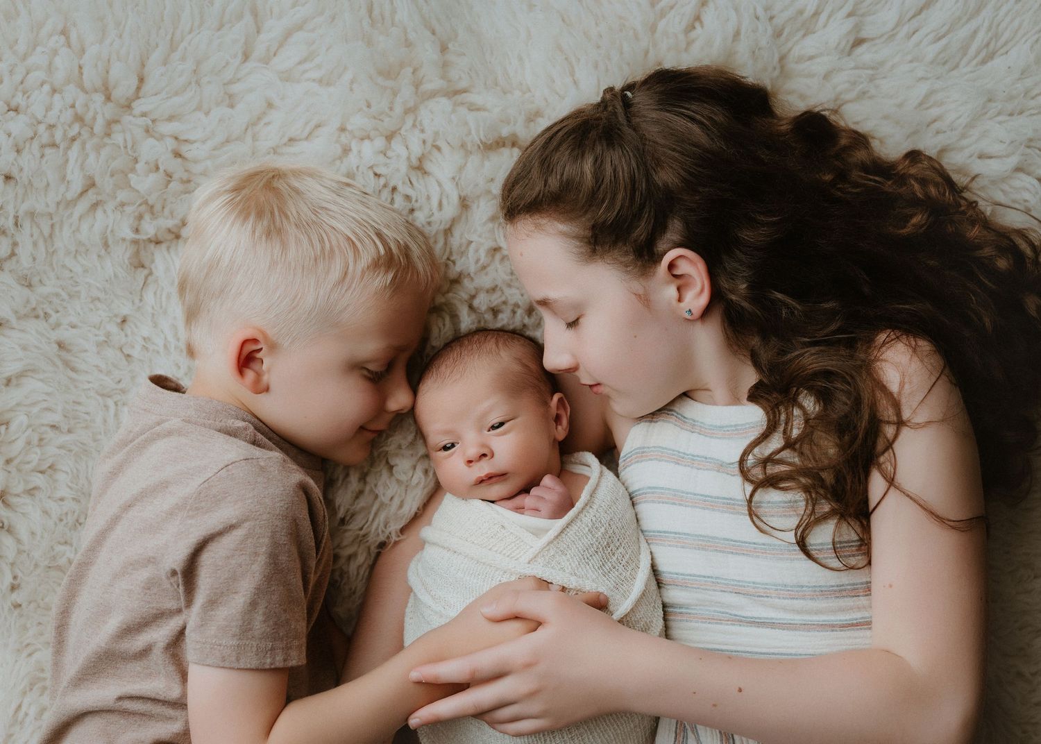 A young boy and mother tenderly cuddle a swaddled newborn baby on a soft white fur rug.