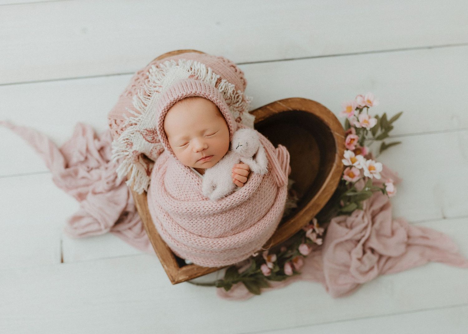 Newborn baby wrapped in pink knit swaddle sleeping in wooden heart bowl with floral bonnet and bunny toy.