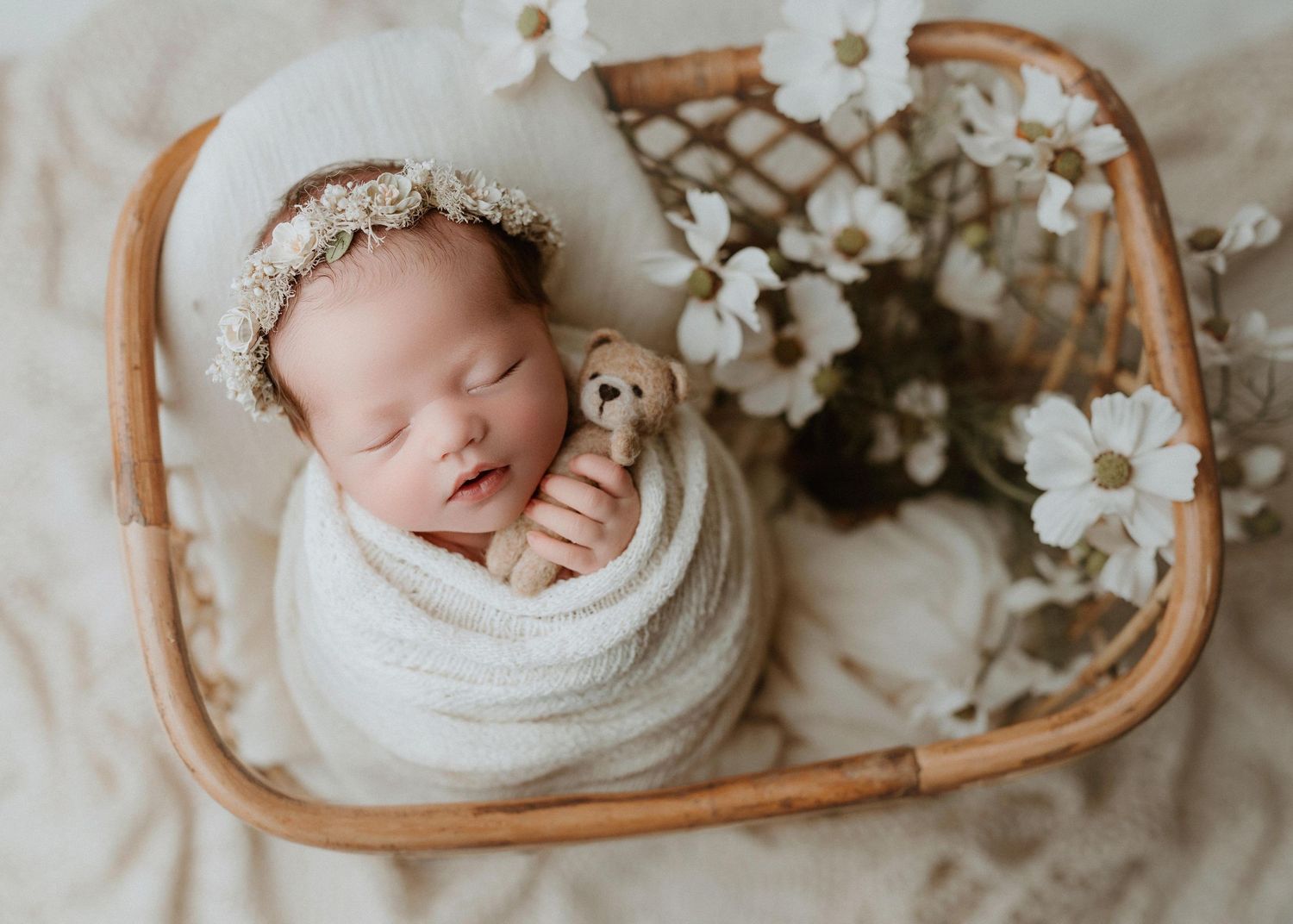 Sleeping newborn baby wrapped in white swaddle holding teddy bear in wicker basket with white flowers.