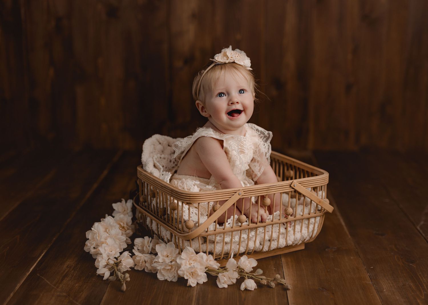 Smiling baby girl in a wicker basket with white flowers, wearing a lace romper and floral headband on a wood floor.