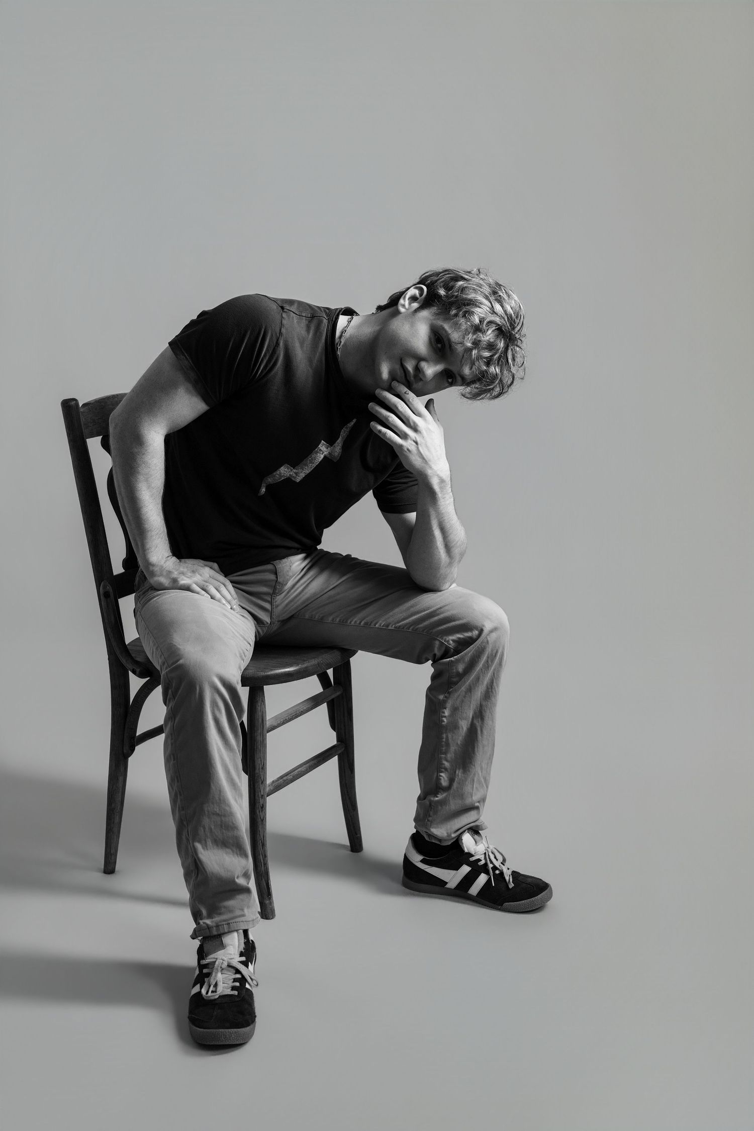 Black and white photo of a young man sitting on a wooden chair, head resting on hand, wearing jeans and a black t-shirt.