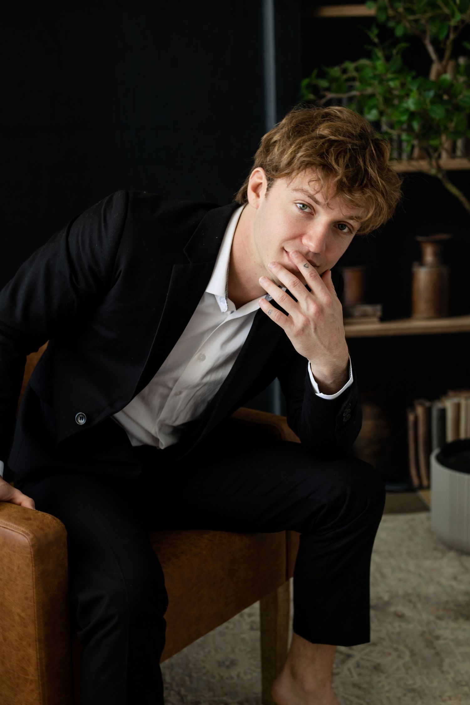 Young man in black suit sitting thoughtfully with hand on chin in a modern dark interior setting.
