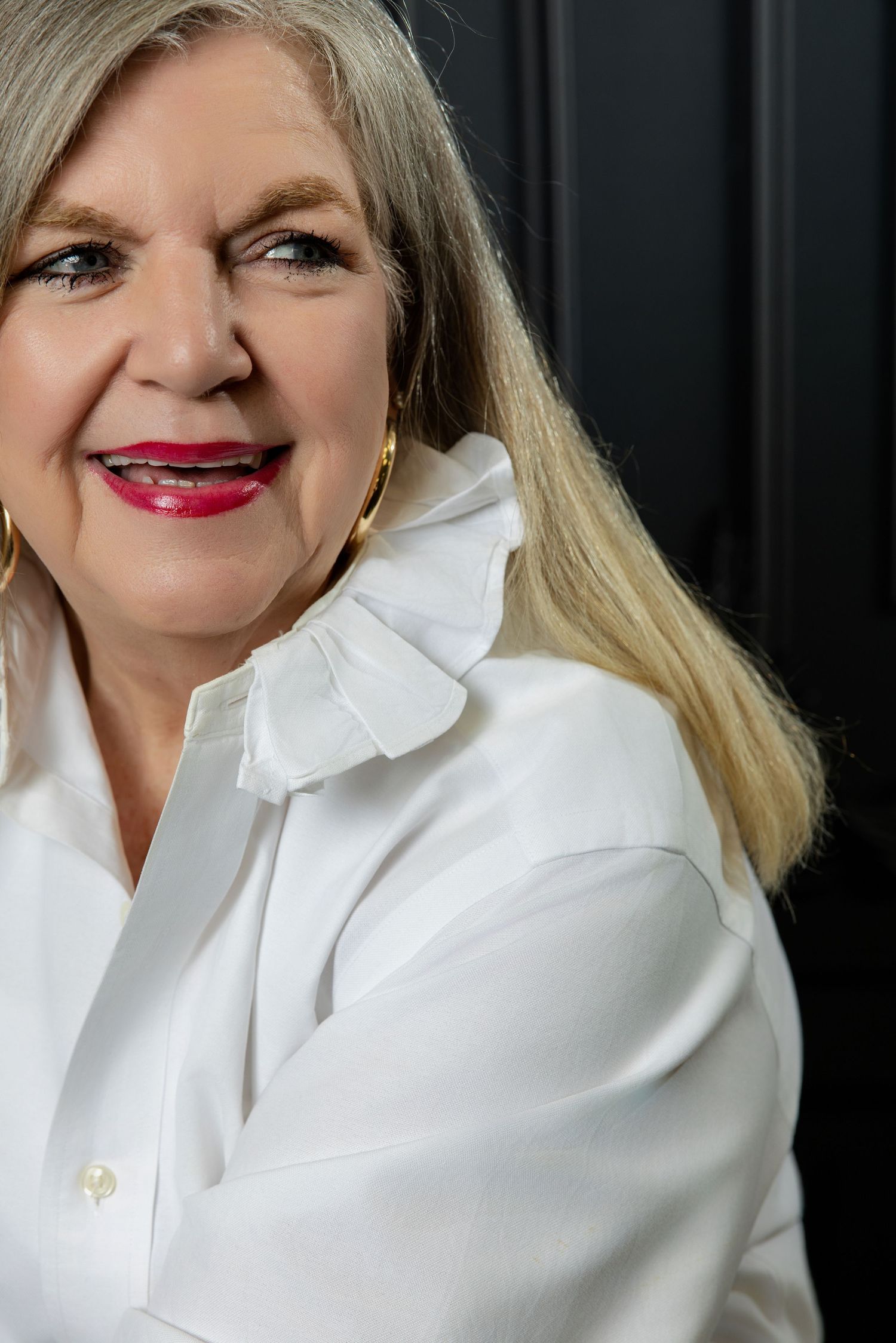 Older woman with blonde hair wearing a white ruffled blouse and gold hoop earrings, smiling confidently.