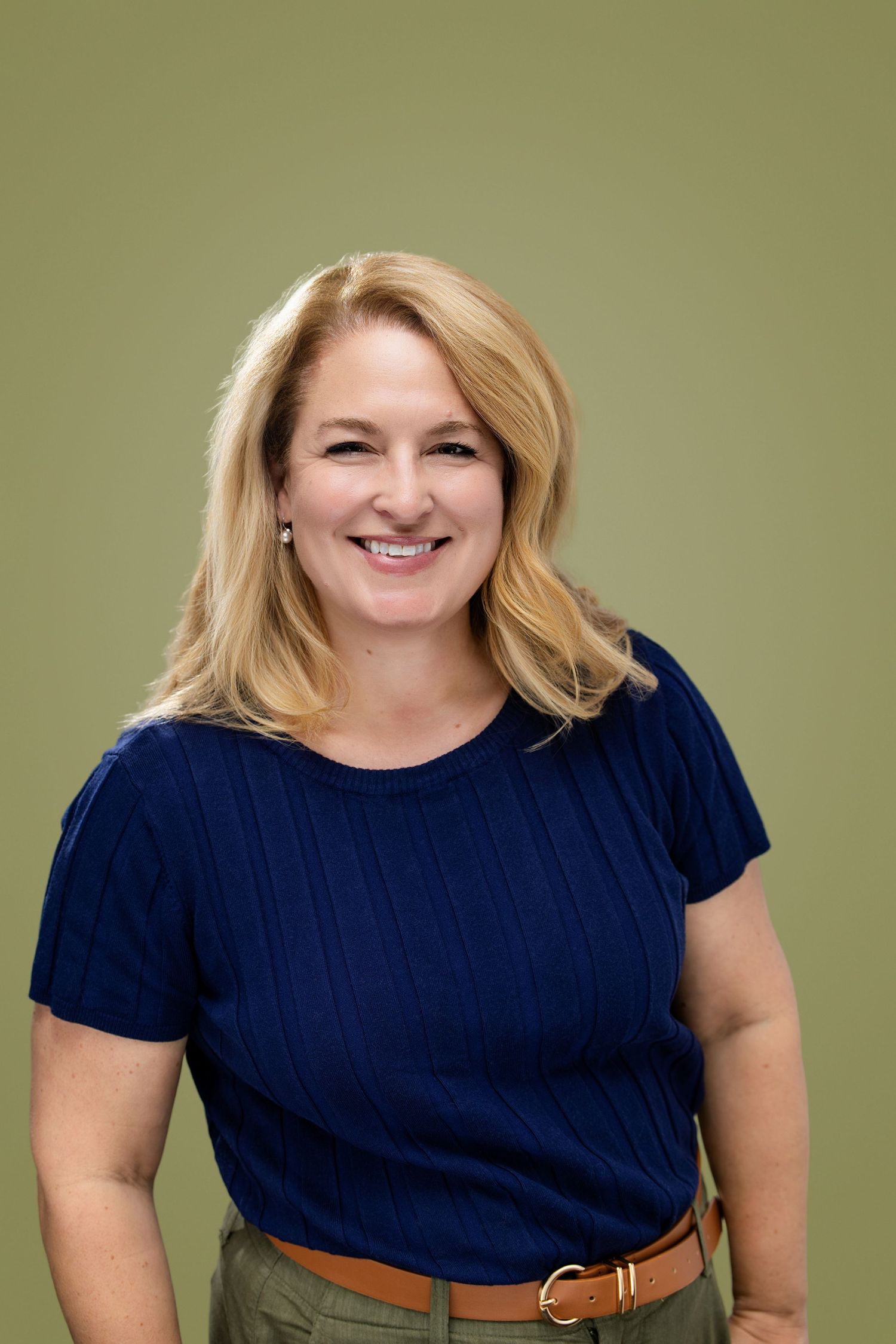 Smiling blonde woman in navy blue top and khaki pants posing against an olive green background.