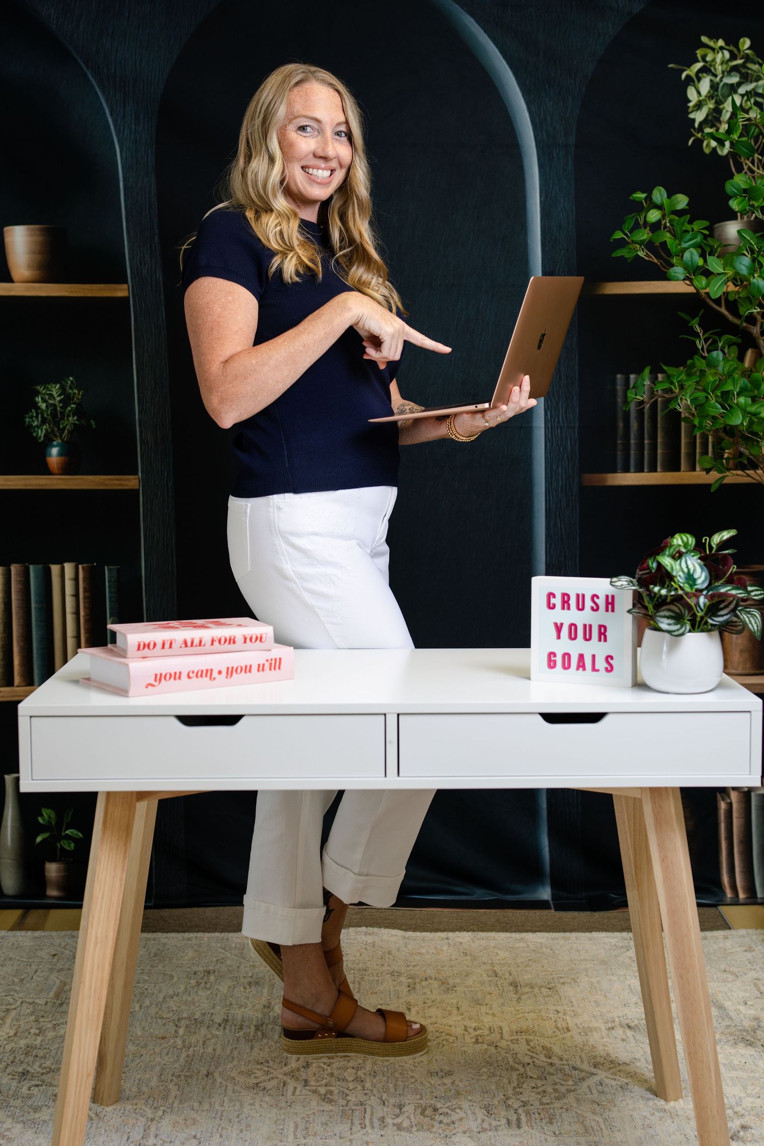 Smiling blonde woman in white pants holding laptop at white desk with motivational books in stylish office.