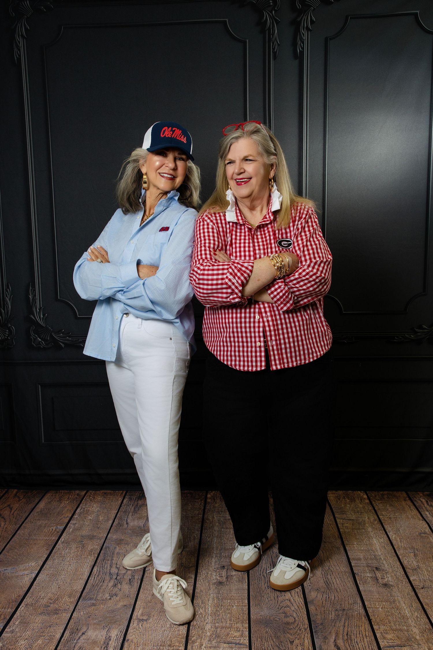 Two smiling women pose back-to-back with arms crossed against a dark wall, wearing casual red and blue outfits.