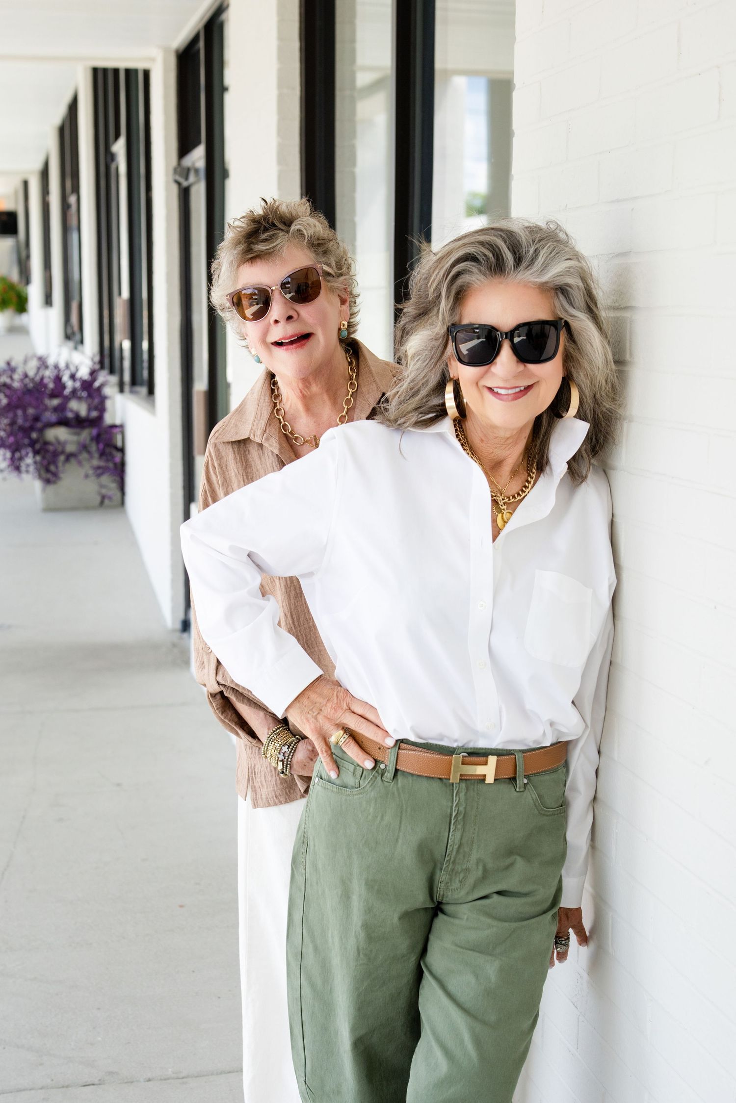 Two stylish older women in sunglasses and gold jewelry pose confidently on a covered walkway.