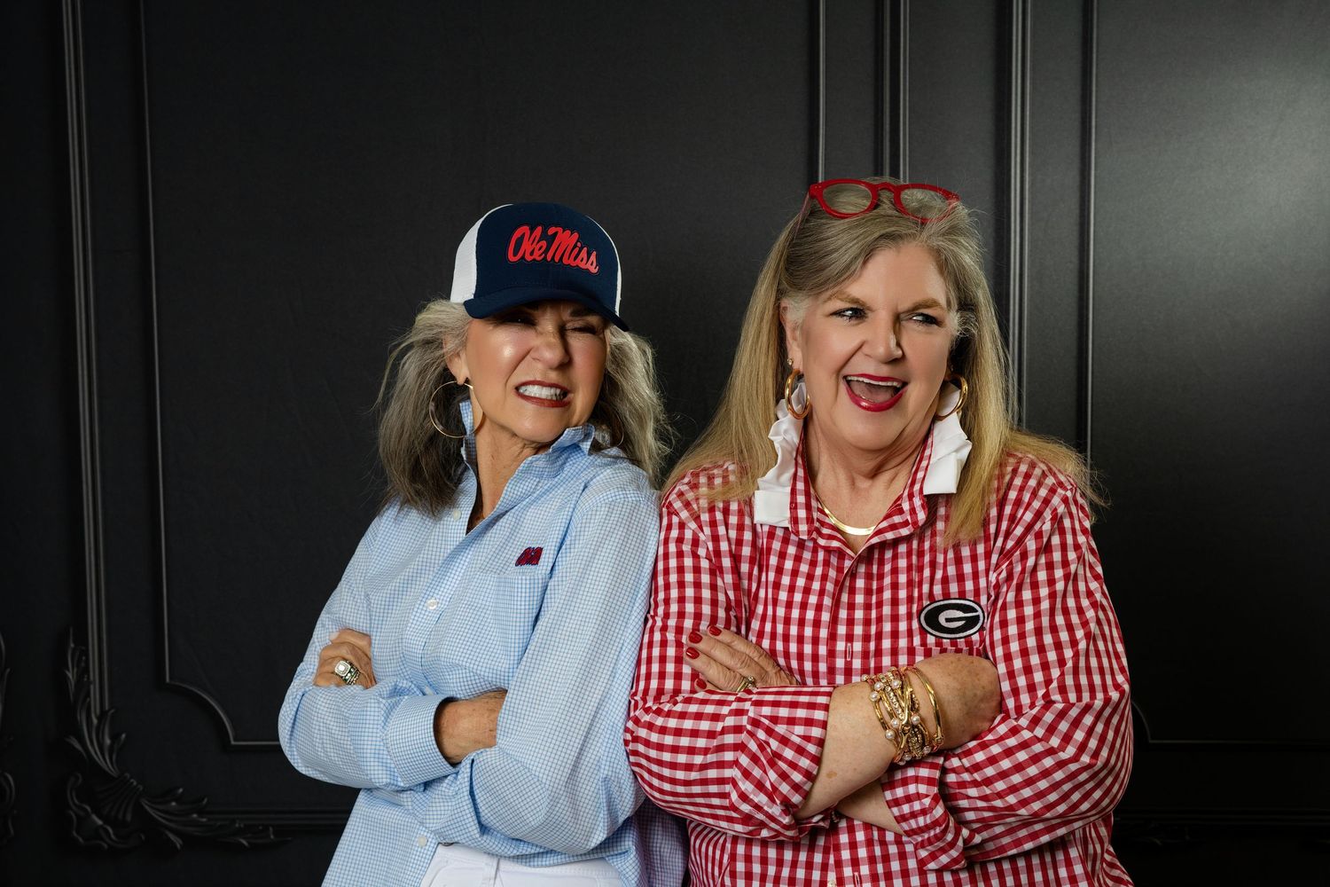 Two women in Ole Miss and Georgia fan gear pose with arms crossed against a dark background.