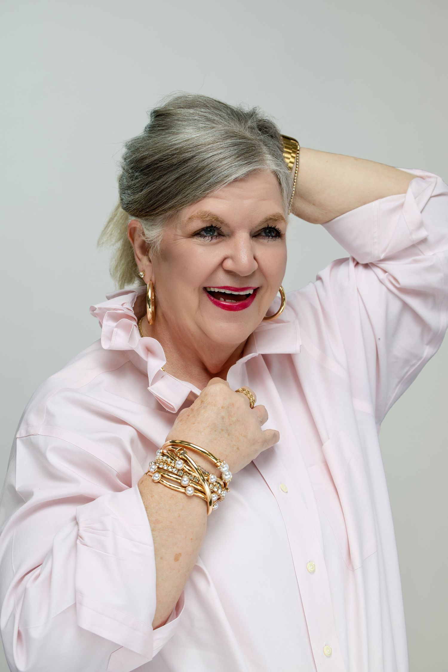 Elegant older woman in pink shirt with gold jewelry, red lipstick, smiling confidently with hand in gray hair.