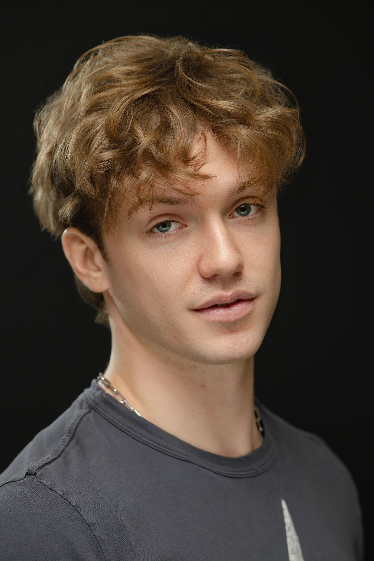 Young man with curly auburn hair and blue eyes wearing a gray t-shirt, posed against a dark background.