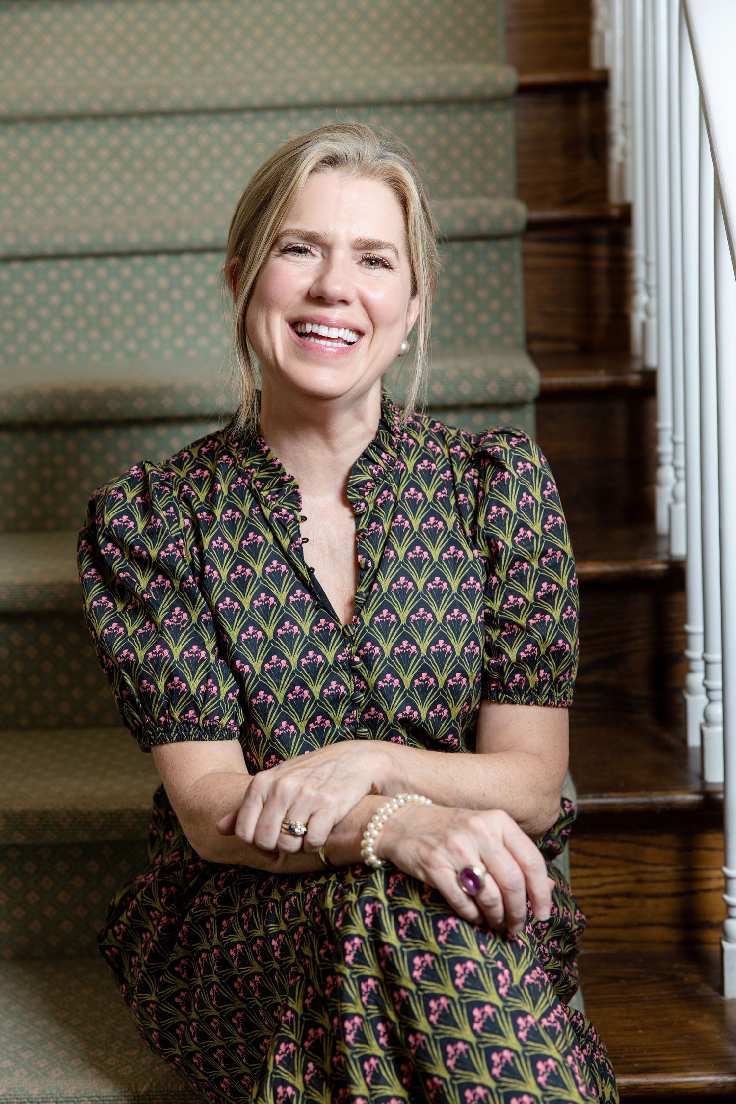 Smiling blonde woman in floral dress sitting on wooden staircase with green carpet runner.