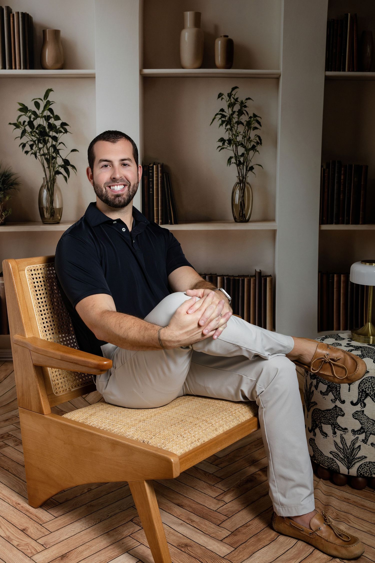 Smiling man in black polo sits cross-legged in wooden chair before bookshelf with vases and plants.