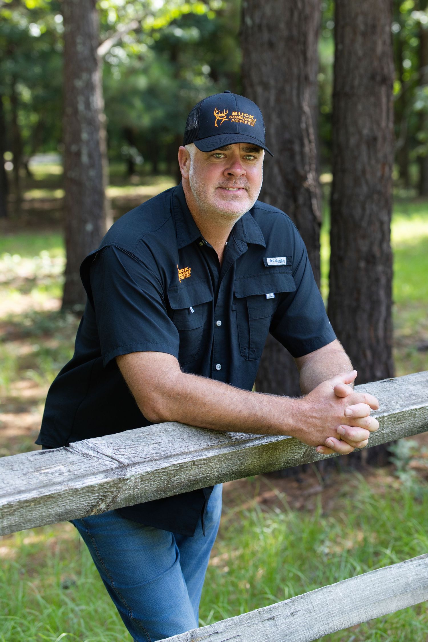 Man in black shirt and cap leans on wooden fence in wooded outdoor setting.