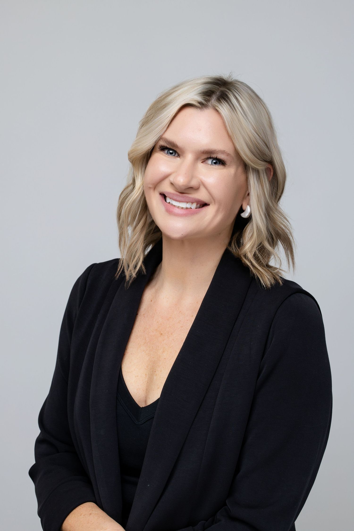 Professional headshot of a smiling blonde woman in a black blazer against a gray background.