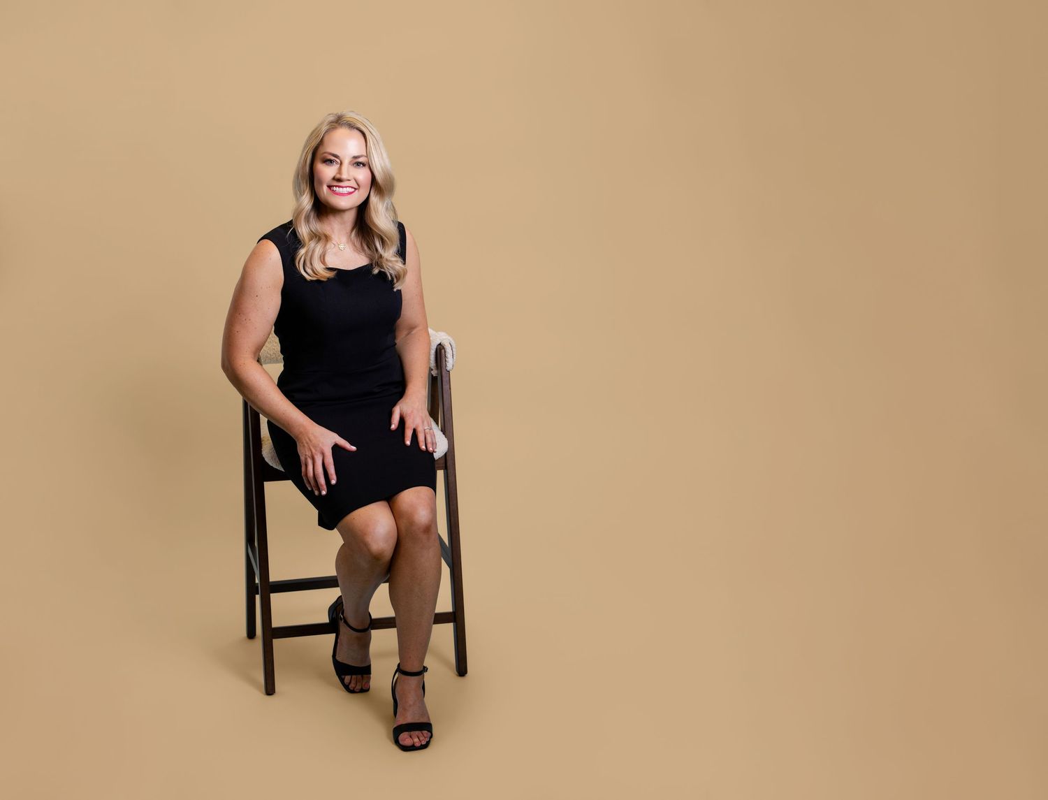 Professional woman in black dress seated on chair, smiling confidently against a warm beige background.