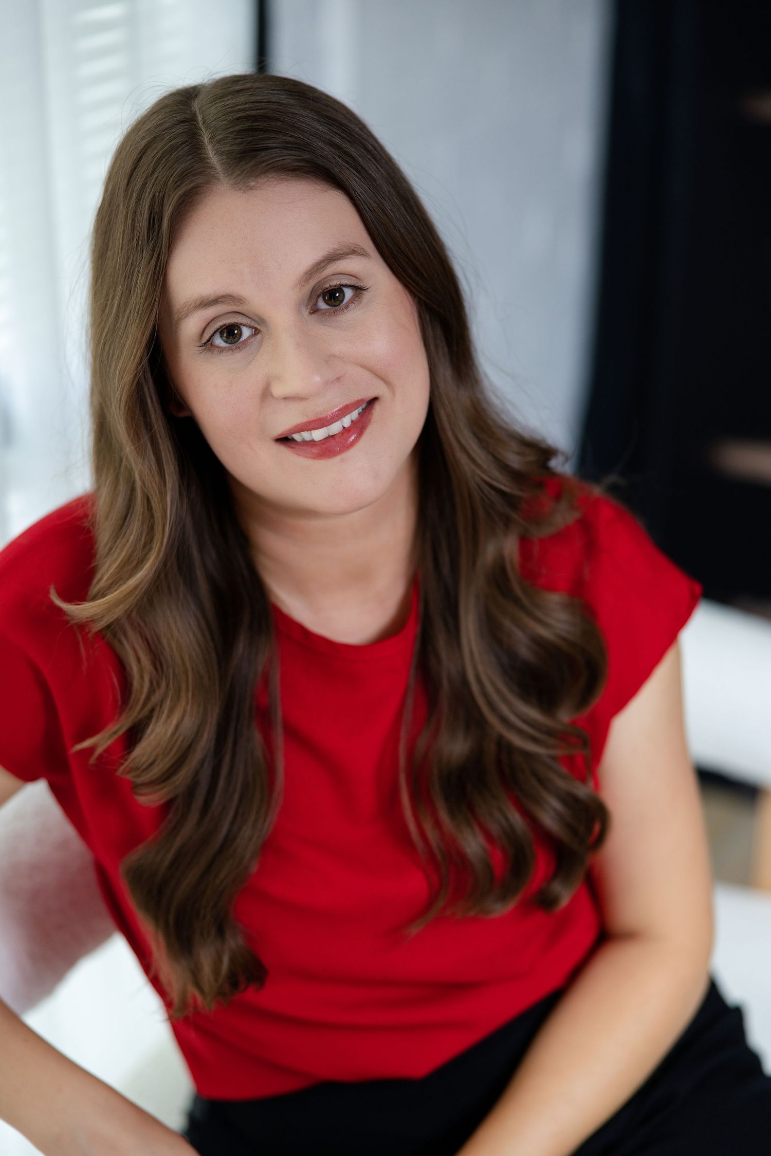 Smiling woman with long brown hair wearing a red sleeveless top, posing confidently indoors.