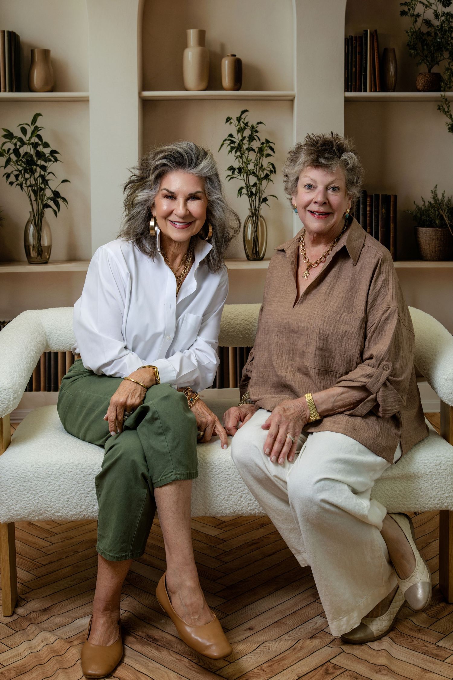 Two elegant older women sitting together on a white chair in a stylish room with shelves and plants.