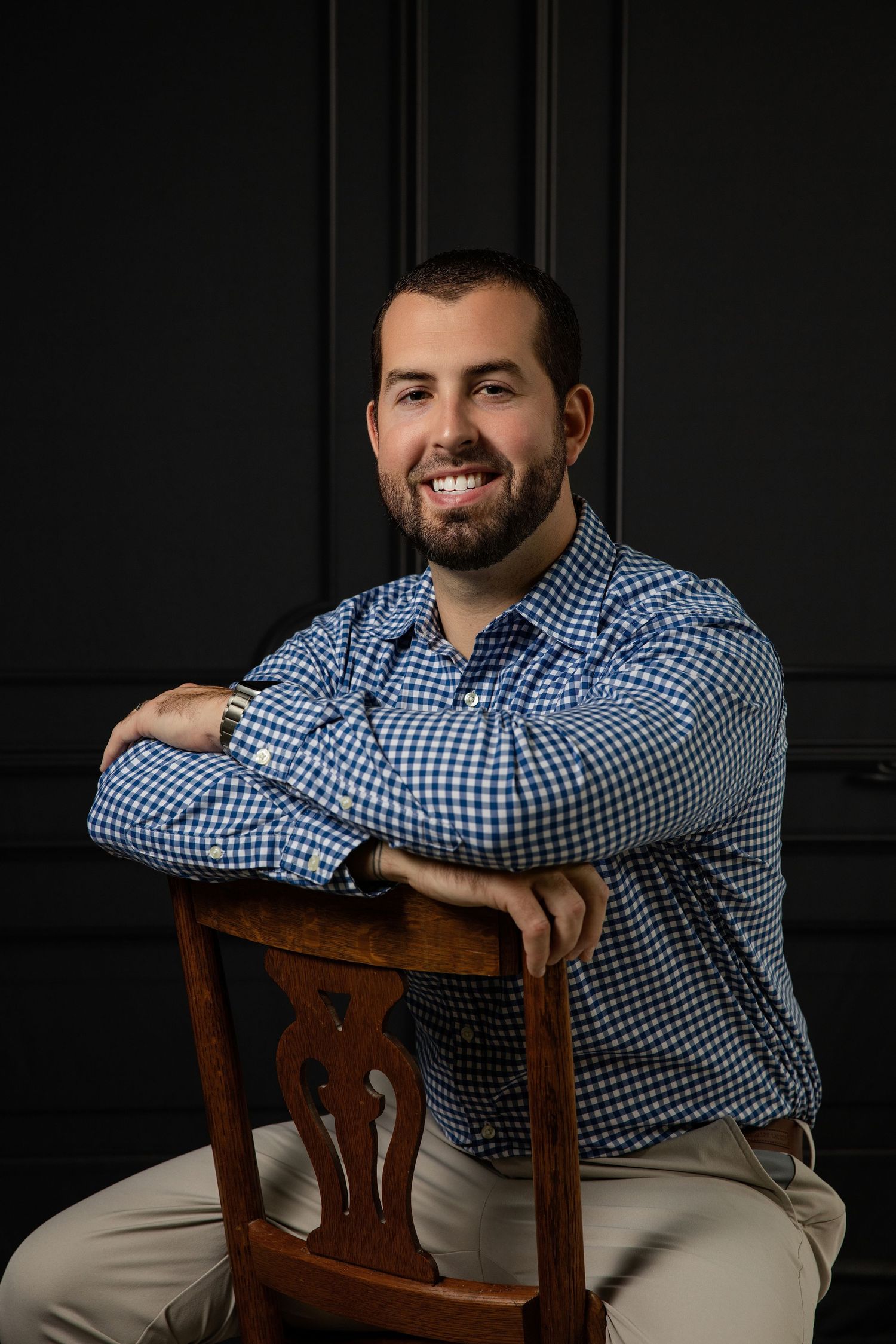 Smiling bearded man in blue gingham shirt sitting relaxed on wooden chair against dark wall.
