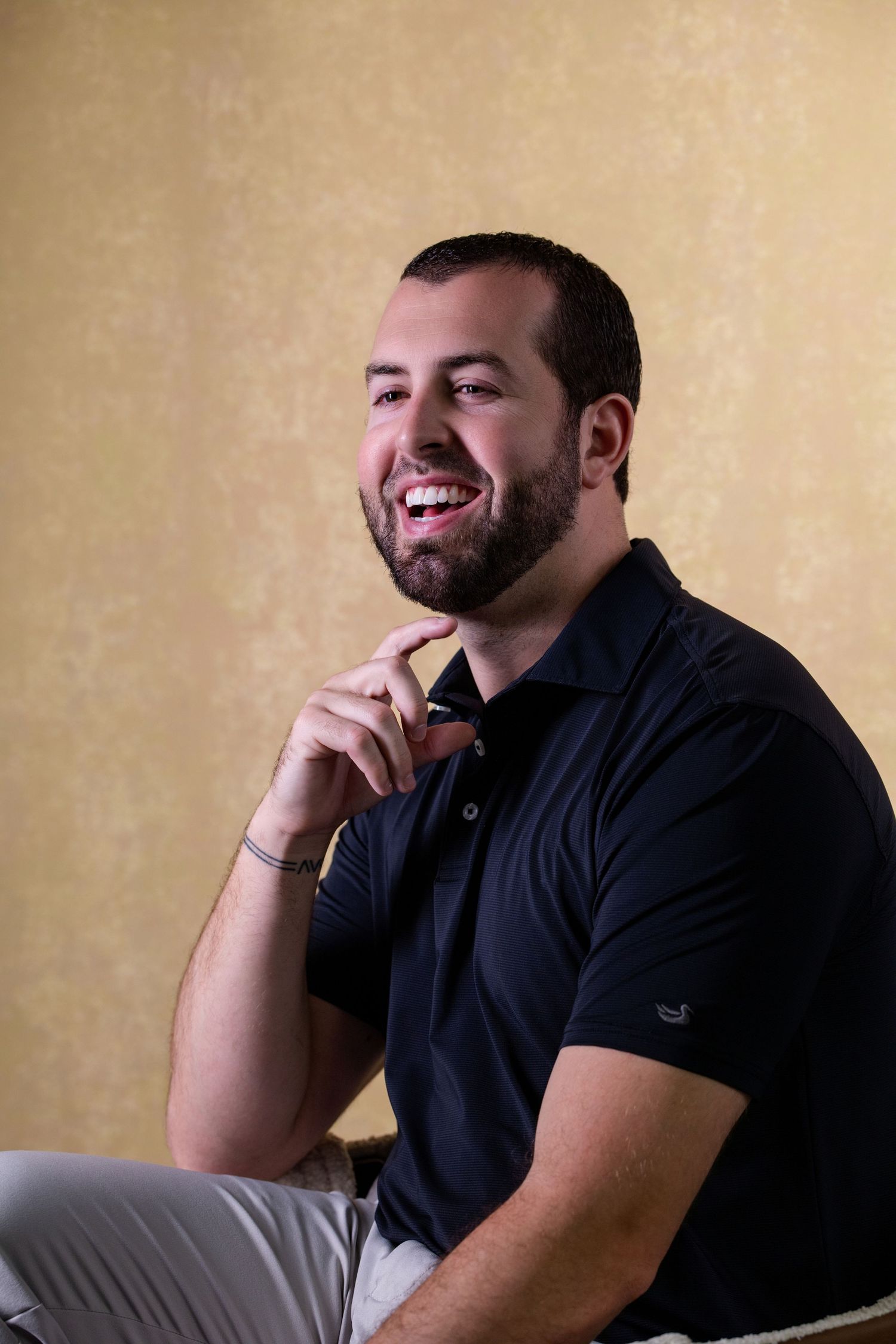 Smiling bearded man in navy polo shirt sitting relaxed with hand on chin against gold wallpaper background.