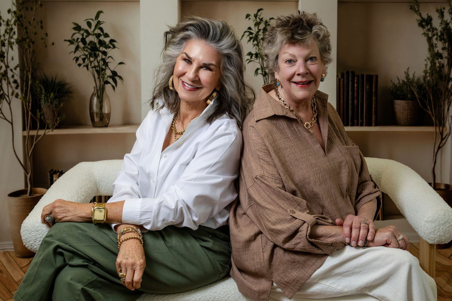 Two elegant older women with gray hair sit back-to-back on a white sofa, smiling warmly in stylish outfits.