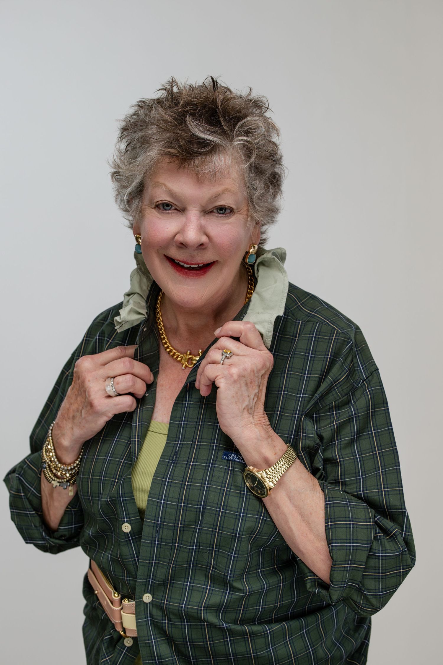 Smiling older woman with short gray hair wearing a green plaid shirt and gold jewelry poses confidently.
