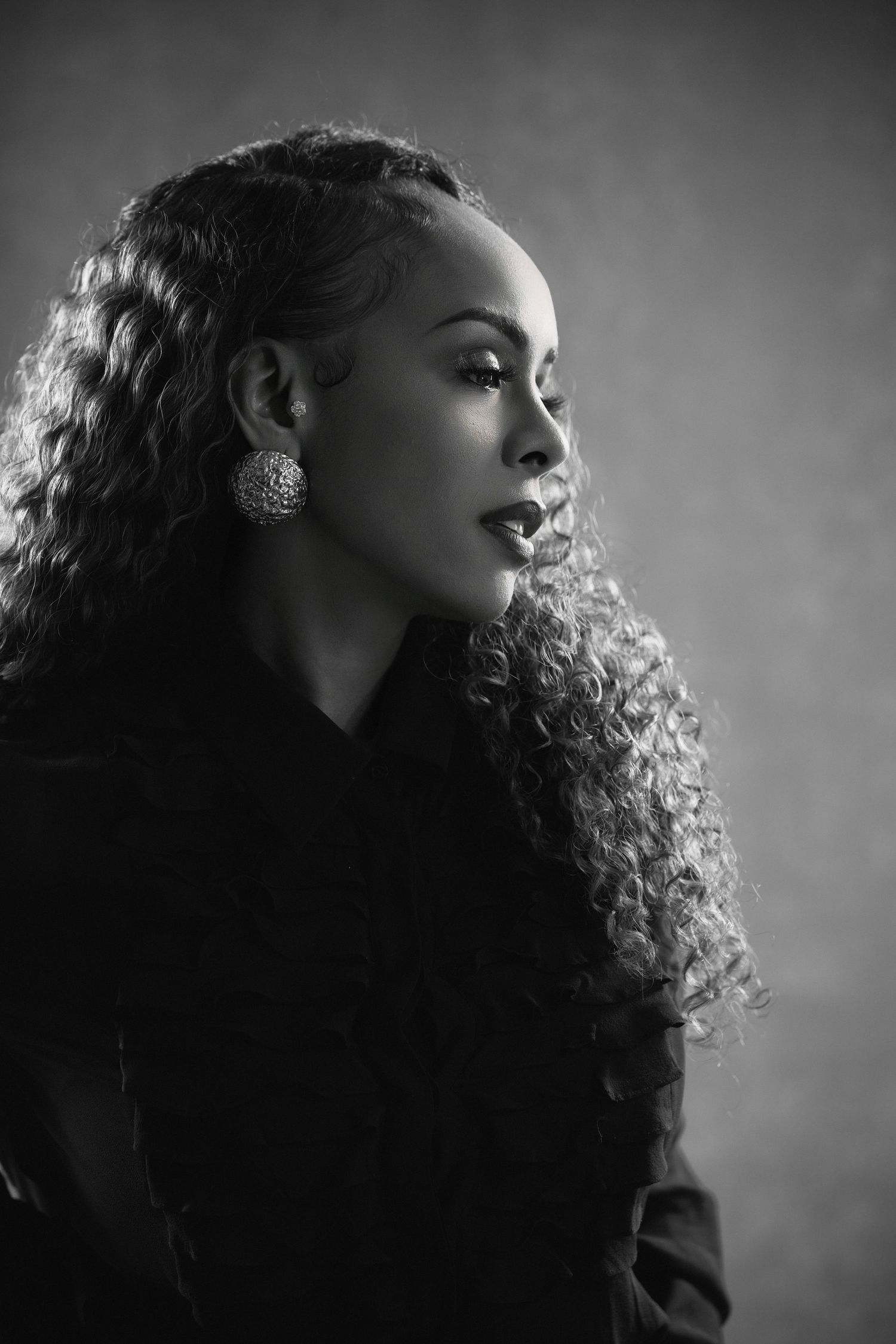 Black and white portrait of a woman with long curly hair wearing large earrings and dark clothing.